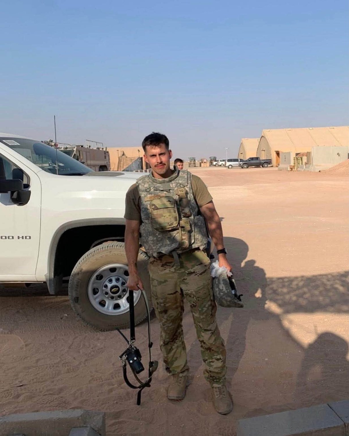 Nick in uniform and gear beside a truck on a desert base