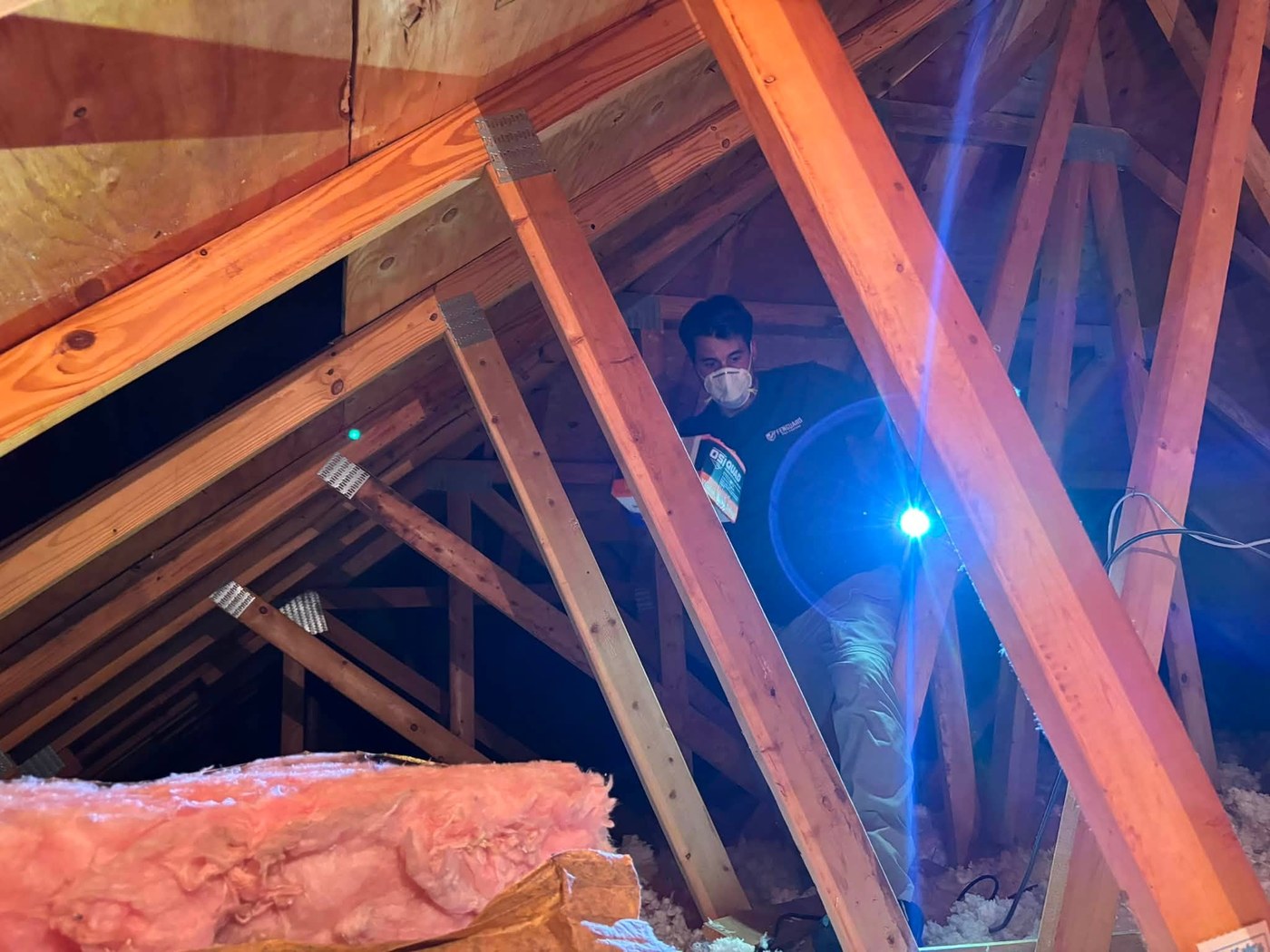 Nick with a respirator and flashlight inspecting attic rafters over pink insulation
