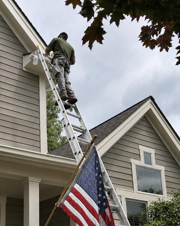 Nick on a ladder at the peak of a house with an American flag on the porch below
