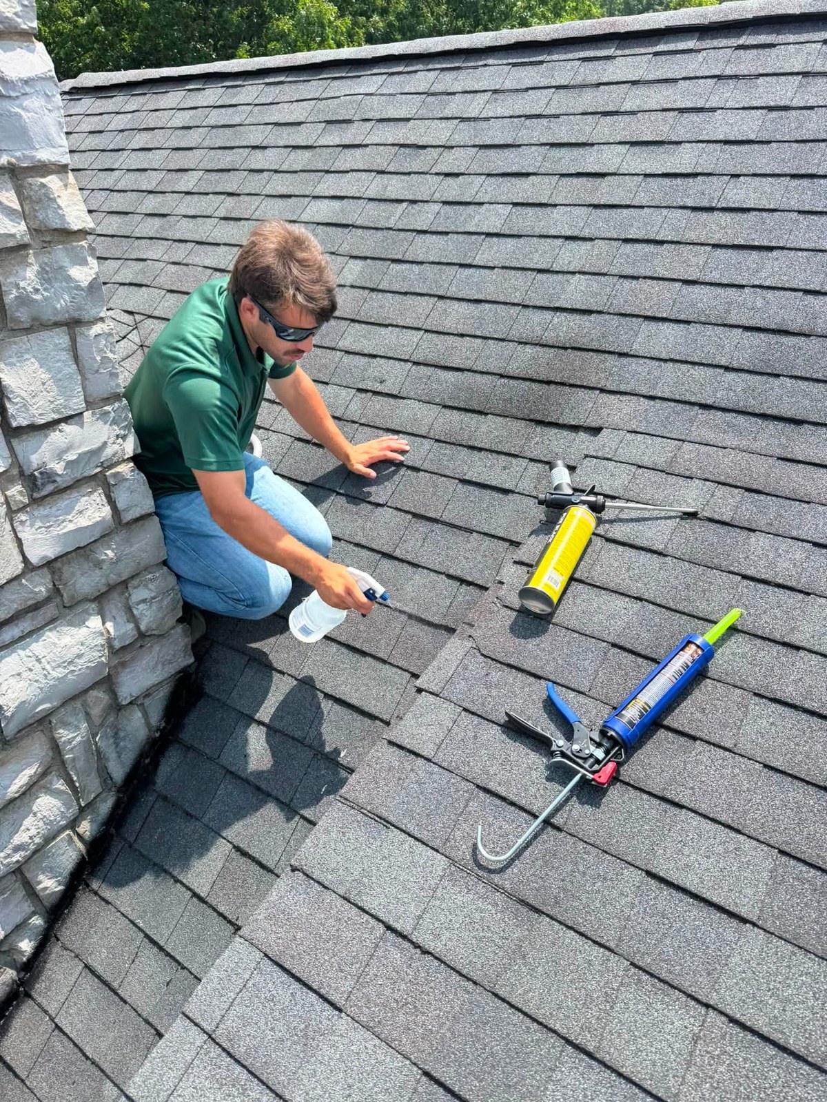 Nick sealing gaps on a shingled roof beside a stone chimney, caulk guns and sealant laid out